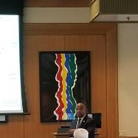 A man stands in front of a podium while 10 seated people listen to him speak